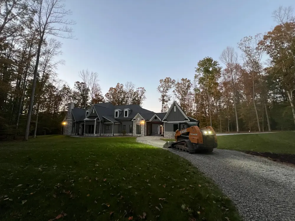 Compact track loader on a new gravel driveway leading to a house in the woods at dusk.